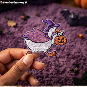 May include: An embroidered patch featuring a white goose wearing a purple witch hat and a purple cape with orange polka dots. The goose is holding an orange jack-o'-lantern pumpkin in its beak. The patch has a distinct purple stitched border.