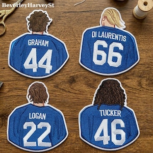 Könnte beinhalten: Vier blau-weiße, gestickte Aufnäher in Form von Trikots. Jeder Aufnäher zeigt einen anderen Namen und eine andere Nummer: Graham 44, Di Laurentis 66, Logan 22 und Tucker 46. Die Aufnäher liegen auf einer Holzoberfläche.