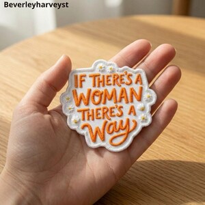 Puede incluir: Un parche bordado blanco con borde festoneado que presenta el texto naranja IF THERE'S A WOMAN THERE'S A WAY. El parche está decorado con pequeñas flores de margarita blancas y amarillas bordadas alrededor del perímetro de las letras.