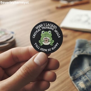 Pode incluir: Um emblema circular preto com a ilustração de um sapo verde segurando uma pequena caneca com um coração. O texto ao redor do sapo diz: "WISH I LACKED CRITICAL THINKING SKILLS Y'ALL SEEM SO HAPPY" em letras maiúsculas brancas.