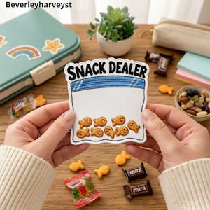 Könnte beinhalten: Ein weißer bestickter Aufnäher mit blauem Reißverschluss-Design und dem schwarzen Text SNACK DEALER. Der Aufnäher zeigt neun kleine orangefarbene fischförmige Cracker, die unten aufgestickt sind. Er wird auf einer Holzoberfläche mit Snacks gehalten.