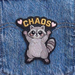 May include: An embroidered patch featuring a grey raccoon with its arms raised, positioned below the word CHAOS in gold thread and two small pink hearts, all on a black background, attached to a blue denim fabric with the text Beverleyharveyst.