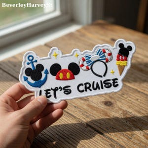 May include: A white embroidered patch with the text "LET'S CRUISE" and various nautical and Mickey Mouse-themed designs. The patch features a blue anchor, Mickey Mouse ears, a bow, and an ice cream cone. The patch is held by a hand.