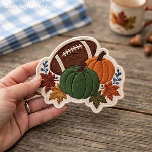 Op de afbeelding: Een geborduurde patch met een bruine football, een groene pompoen, een oranje pompoen en diverse herfstbladeren in bruine, oranje en groene tinten. De patch heeft een crèmekleurige achtergrond en een geschulpte rand met gedetailleerd stikwerk.