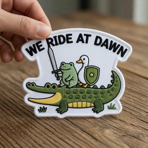 May include: Embroidered patch featuring a green alligator with a frog holding a sword and a duck with a shield. The patch has the text "WE RIDE AT DAWN" in black on a white background. The patch is held up by a hand.