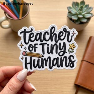 May include: A white embroidered patch with a scalloped edge featuring the black text "Teacher of Tiny Humans" in a cursive font. The design includes a yellow pencil, a small yellow flower with green leaves, and several small four-pointed stars.