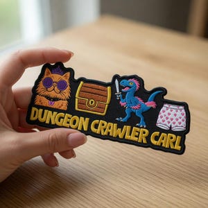 Könnte beinhalten: Schwarzer, gestickter Aufnäher mit dem Text "DUNGEON CRAWLER CARL". Der Aufnäher zeigt eine Katze mit Sonnenbrille und Krone, eine Schatztruhe, einen blauen Dinosaurier mit rosa Irokesenschnitt, der ein Schwert hält, und eine Unterhose mit Herzen.