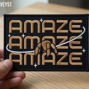 May include: Black embroidered patch featuring the word "AMAZE" in gold, repeated three times. A white curved line and stars are also embroidered. The patch has a black border.