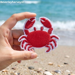 Peut inclure: Un écusson brodé en forme de crabe rouge avec une bordure blanche. Le crabe présente deux pinces levées, deux petits yeux et huit pattes. Le motif est réalisé avec une texture de fil dense, créant un aspect rouge vif sur un contour blanc.