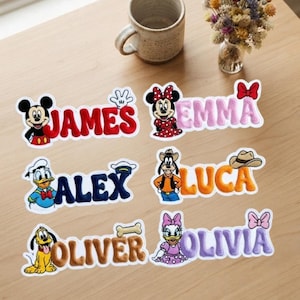 Könnte beinhalten: Sechs bestickte Namensaufnäher mit Disney-Figuren. Die Aufnäher zeigen die Namen James in Rot, Emma in Pink, Alex in Dunkelblau, Luca in Orange, Oliver in Braun und Olivia in Lila, jeweils ergänzt durch ein passendes Figuren-Symbol.