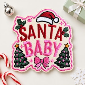 May include: An embroidered patch featuring the text SANTA BABY in red and pink thread. The design includes a red Santa hat, two green Christmas trees with pink ornaments, holly leaves with red berries, and a pink bow on a cream background.