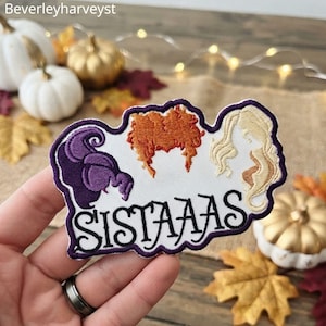 May include: An embroidered patch featuring three stylized hair silhouettes in purple, orange, and blonde, positioned above the black embroidered text "SISTAAAS" on a white background with a dark purple border outline.