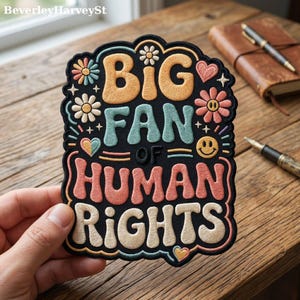 May include: Embroidered patch with the text "BIG FAN OF HUMAN RIGHTS" in a retro font. The patch features a black background with colorful floral and heart accents. The patch is held by a hand.