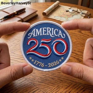 May include: A round blue patch with the word "AMERICA" in white arched text. The number "250" is in red and blue, with the years "1776-2026" below. The patch has a white border and is held by two hands.