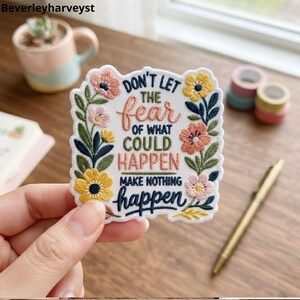 Könnte beinhalten: Ein weißer bestickter Aufnäher mit einem Blumenrand aus rosa, gelbem und grünem Garn. Der Text lautet: Don't let the fear of what could happen make nothing happen, gestickt in dunkelblauem, pfirsichfarbenem und olivgrünem Garn.