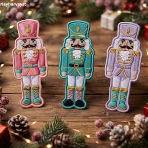 Puede incluir: Tres parches bordados de cascanueces en rosa, verde azulado y lavanda. Cada figura lleva un sombrero alto, un uniforme detallado con detalles dorados y pantalones blancos. Los parches presentan un bigote, una barba blanca y botas. Texto: Beverleyharveyst.
