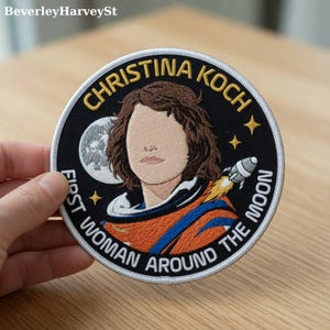 Op de afbeelding: Geborduurde patch met Christina Koch, met de tekst "CHRISTINA KOCH" en "FIRST WOMAN AROUND THE MOON". De patch heeft een zwarte achtergrond met een portret, een raket, de maan en sterren.