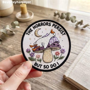 Puede incluir: Un parche bordado circular con fondo blanco y borde negro. Presenta una oca blanca sobre un hongo morado, una luna creciente y flores pequeñas. El texto dice THE HORRORS PERSIST BUT SO DO I.