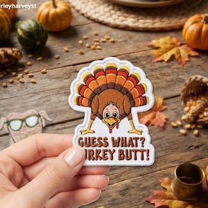Könnte beinhalten: Ein bestickter Aufnäher mit einem Cartoon-Truthahn, der sich bückt und seine Schwanzfedern auffächert. Der Text unter dem Truthahn lautet "GUESS WHAT? TURKEY BUTT!". Der Aufnäher hat einen weißen Rand und wird vor einer rustikalen Holzoberfläche gehalten.