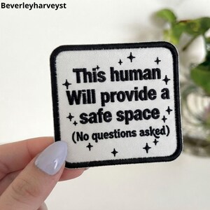 May include: A square white embroidered patch with a thick black border. The text reads "This human Will provide a safe space (No questions asked)" in black lettering, surrounded by small black star icons scattered across the white background.