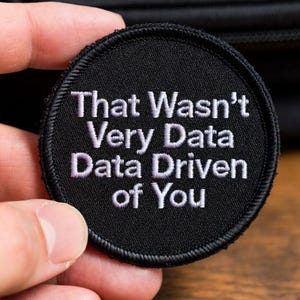 May include: A circular black embroidered patch with a stitched border. The patch features white text that reads, "That Wasn't Very Data Data Driven of You," arranged in five lines across the center of the circular fabric surface.