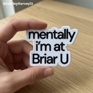 Puede incluir: Parche blanco con texto negro "mentally I'm at Briar U". El parche tiene un borde blanco y está sujeto por una mano. El parche está sobre una superficie de madera marrón claro.