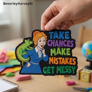 May include: Embroidered patch with a cartoon woman in a blue dress and a green lizard. The patch reads "TAKE CHANCES MAKE MISTAKES GET MESSY" in multiple colours. The patch is black with a white border.