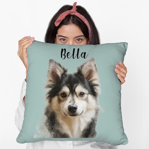 May include: A square, light blue pillow featuring a portrait of a dog with black, white, and tan fur. The dog's name, "Bella," is written in black script across the top of the pillow.
