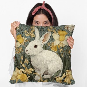 May include: Decorative square pillow featuring a white rabbit illustration amidst yellow daffodils and green foliage against a teal background. The design has a vintage, artistic style, suitable for home decor.
