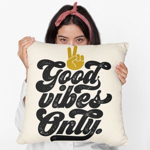 Pode incluir: Uma almofada branca com o texto "Good vibes only." em letras pretas com um sinal de paz dourado acima do texto.