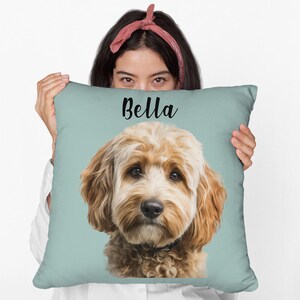 May include: A square, light blue pillow featuring a portrait of a dog with tan and white fur. The dog's name, "Bella", is written in black script across the top. The pillow is held up by a person.