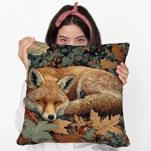May include: A decorative pillow featuring a sleeping fox illustration. The fox is depicted in shades of brown and orange, nestled among autumn leaves. The pillow's background showcases a floral and foliage pattern in green and gold.