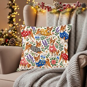 May include: A square throw pillow featuring a repeating pattern of Basenji dogs wearing various colorful sweaters, including red, yellow, and blue designs with heart and floral motifs, interspersed with illustrations of lavender and daisies.