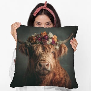 May include: A decorative pillow featuring a Highland cow with long, reddish-brown fur and a floral crown. The cow has large horns and is centered on a dark background. The pillow is held by a person.