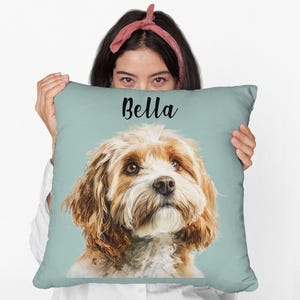 Op de afbeelding: Een vierkant sierkussen met een lichtblauwe achtergrond met een gedetailleerd portret van een hond met bruine en witte vacht. De naam "Bella" staat in zwart schrift bovenaan.