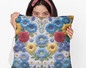 Könnte beinhalten: Ein dekoratives Kissen mit einem lebendigen Blumenmuster. Das Kissen zeigt ein Muster aus blauen, gelben und roten Blumen, darunter Gänseblümchen und Rosen, vor einem weißen Hintergrund. Das Design hat eine strukturierte, dreidimensionale Optik.