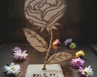 May include: Wooden rose with a brown stem and leaves, standing on a wooden base with the word "mom" carved into it. The rose is surrounded by small white and pink flowers.