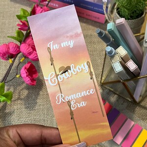May include: A pink and orange bookmark with a sunset background. The bookmark has white text that reads "In my Cowboy Romance Era".