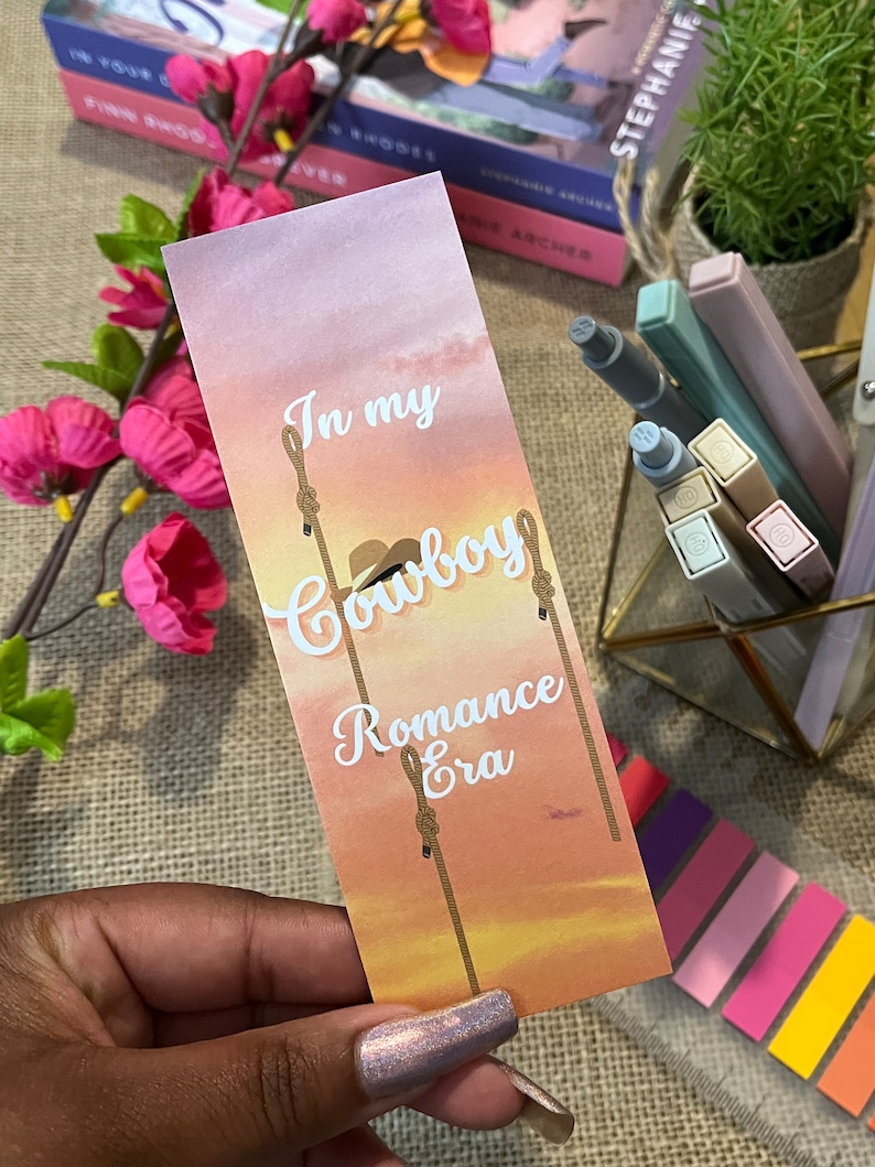 May include: A pink and orange bookmark with a sunset background. The bookmark has white text that reads "In my Cowboy Romance Era".