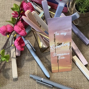 May include: A close-up of a bookmark with the text "In my Cowboy Romance Era" on a pink and purple gradient background. The bookmark is surrounded by a variety of pastel colored pens and markers.
