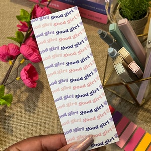May include: A white bookmark with the phrase "good girl" repeated in pink and purple lettering. The bookmark is being held by a hand with a glittery nail polish.