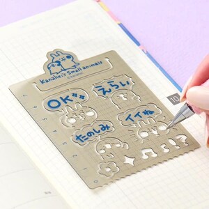 May include: A metallic stencil featuring Kanahei's Small Animals branding, with cutouts for speech bubbles containing the text OK, Japanese characters, and various icons including a flower, music note, and exclamation point, with a 7-inch ruler edge.