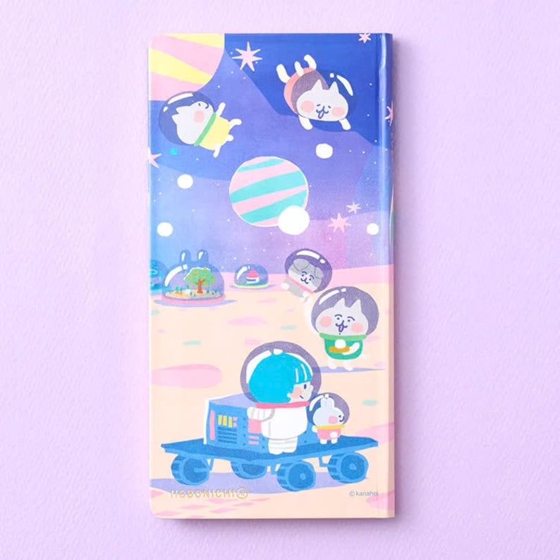 May include: A rectangular notebook with a space-themed cover featuring cartoon characters in astronaut helmets, a blue lunar rover, planets, and stars against a gradient purple and peach background. The text Hobonichi is printed at the bottom.
