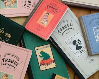 May include: A collection of various colored "Travel Notebook" journals. The notebooks feature illustrations and text, including "Travel Notebook" and "Demestore Design". Colors include pink, red, green, gray, blue, and tan. Some notebooks have illustrations of a child with a dog, and a frog.