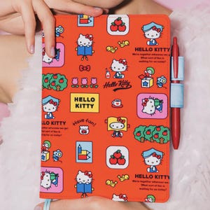 Puede incluir: Un cuaderno naranja con un patrón repetitivo de Hello Kitty que incluye manzanas, libros y ratones. El texto dice Hello Kitty, Have fun!, y We're together wherever we go! What sort of fun is waiting for us today? Un bolígrafo rojo está sujeto al lateral.