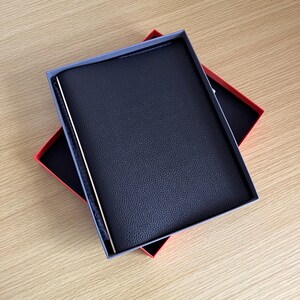 May include: A black leather notebook with a gold trim is displayed in a red box with a black interior.