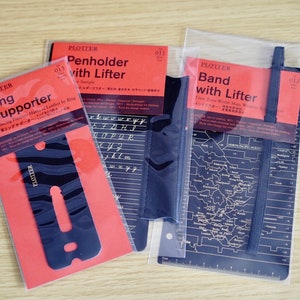 Può includere: Tre accessori in pelle nera per planner in un imballaggio di plastica trasparente. Gli accessori sono un supporto per anelli, un portapenne con sollevatore e una fascia con sollevatore. La confezione include il nome del prodotto e una descrizione del prodotto.