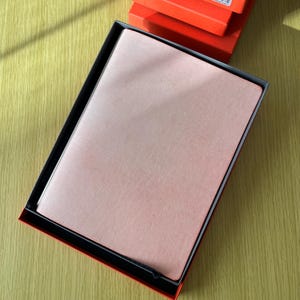 May include: A rectangular notebook with a textured, light pink cover is nestled inside a black-lined, bright orange presentation box. The notebook is positioned centrally within the box, which is set against a light-colored wooden surface.