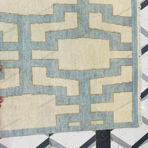 Elegant handwoven wool rug photographed from a corner angle to display its precise geometric pattern and neat edges. This modern flatweave rug features a minimalist abstract design in neutral beige with soft blue accents. Ideal as a living room rug or bedroom rug, it complements modern, bohemian, and coastal décor styles. Crafted by skilled artisans, this handmade area rug offers durability, style, and timeless appeal for contemporary homes.