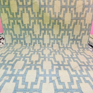 Room scene featuring a large handmade wool rug placed on a patterned floor, showcasing its modern geometric design. The calming blue and beige tones enhance the overall interior aesthetic. This flatweave area rug is ideal for contemporary homes, offering durability, comfort, and style. Perfect for living rooms or bedrooms, it blends handcrafted quality with modern home décor trends.